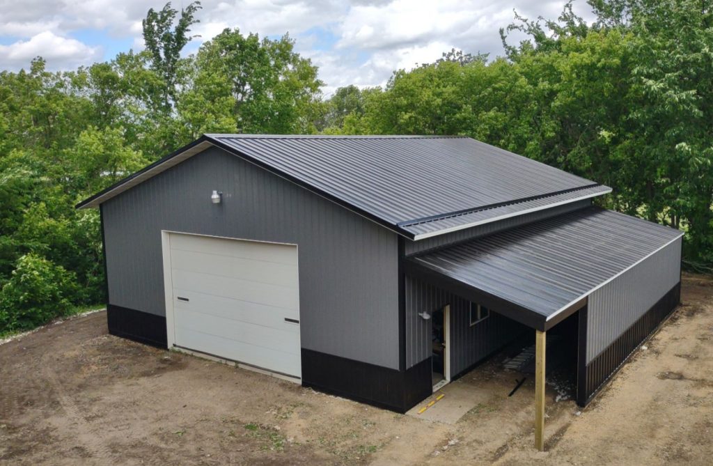 Gray metal garage with attached carport surrounded by trees